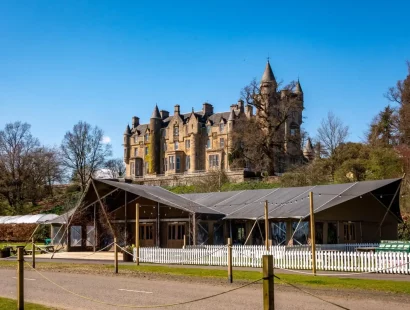 The photo shows a beautiful castle-like building against the backdrop of a bright blue sky. The building is large and has turrets. In the foreground, there is a green marquee.